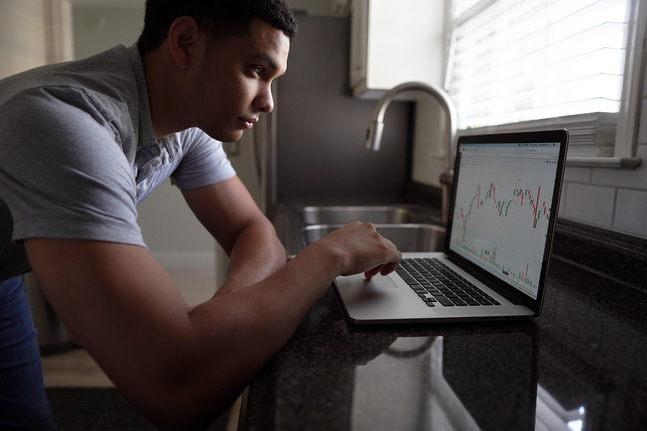 Jeune homme concentré analysant un graphique boursier sur l’écran de son ordinateur portable dans une cuisine.